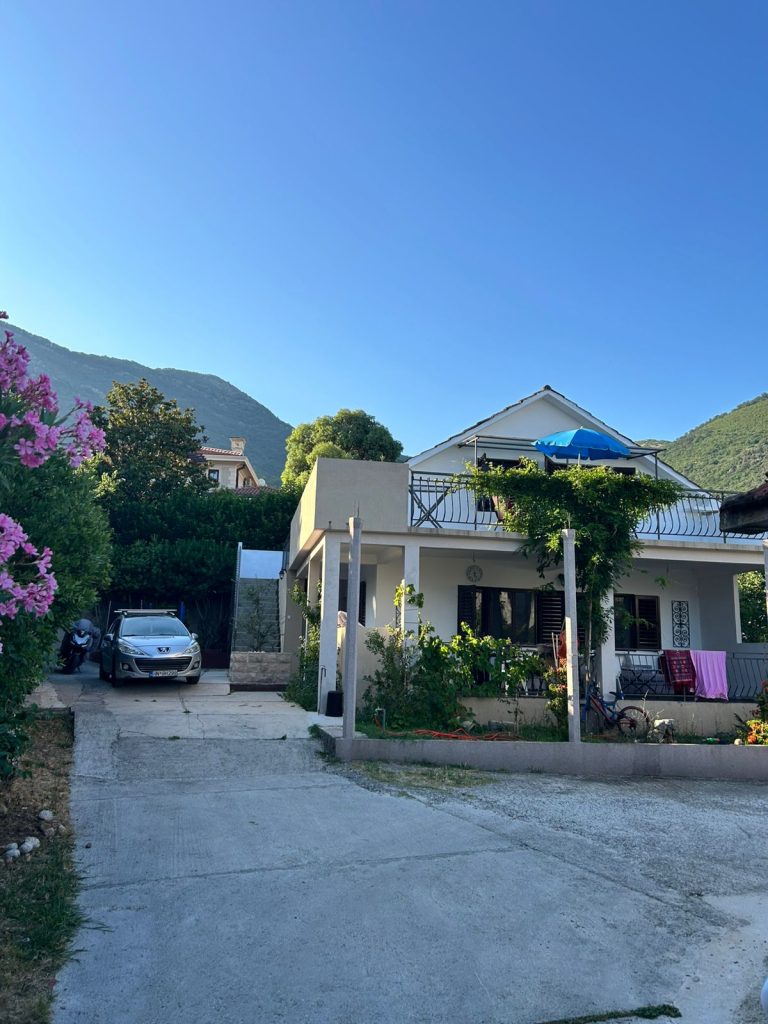Kuća sa apartmanima u Baošićima, Crna Gora, okružena zelenilom i planinama u pozadini. Na slici se vidi dvorište sa betonskim prilazom, parkiranim automobilom i motorom, terasa sa plavom tendom i sušenjem veša. Ova kuća nudi smeštaj u mirnom delu Baošića, blizu obale i plaže, sa obezbeđenim parking mestom.
